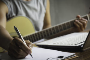 Composer holding pencil and writing lyrics in paper. Musician playing acoustic guitar.